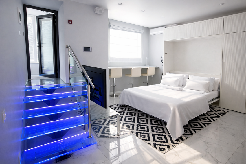 Раннее утро в апартаментах. Аккуратно заправленная кровать, мягкий свет из окна и стеклянной входной группы. Стеклянная лестница с RGB-подсветкой в глубоком ярко-синем оттенке создаёт брутальную, спокойную атмосферу.  При открытых жалюзи пространство напо